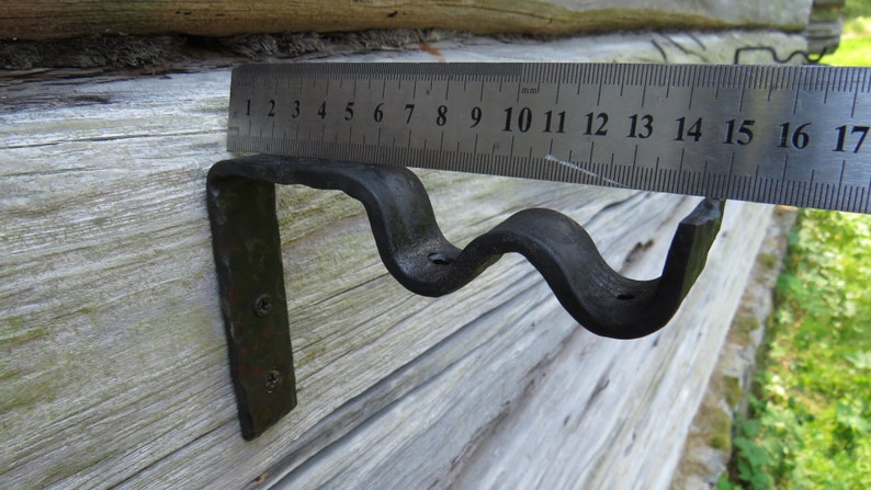 Op de afbeelding: Een zwarte metalen beugel met een golvend ontwerp, gemeten met een liniaal die in centimeters is gemarkeerd. De beugel is bevestigd aan een houten oppervlak.