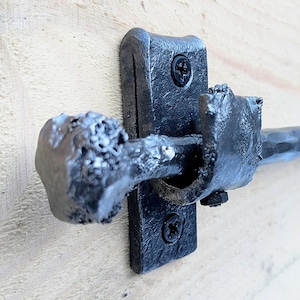 Könnte beinhalten: Nahaufnahme eines schwarzen, handgeschmiedeten Metallverschlusses. Der Verschluss hat einen strukturierten, abgerundeten Griff und eine rechteckige Rückplatte mit zwei Schrauben. Das Metall hat ein gehämmertes Aussehen, was auf einen rustikalen, handwerklichen Stil hindeutet.