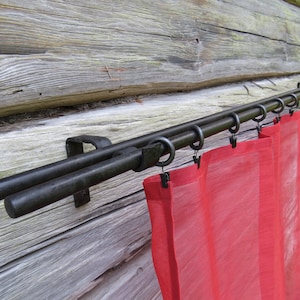 Op de afbeelding: Dubbele zwarte metalen gordijnroede met rode, doorzichtige gordijnen die eraan hangen. De roede is bevestigd aan een houten muur.