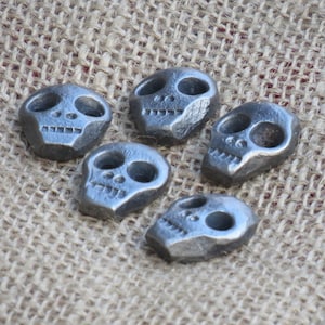 Puede incluir: Cinco pequeños amuletos de metal en forma de calavera de color plateado. Las calaveras tienen un diseño sencillo con dos cuencas de los ojos, una nariz y una boca.