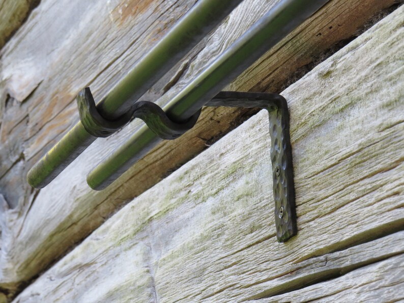 Op de afbeelding: Zwarte metalen dubbele gordijnroedebeugels gemonteerd op een houten muur. De beugels hebben een gehamerde afwerking en zijn ontworpen om twee roedes te dragen.