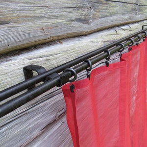 Op de afbeelding: Dubbele gordijnroede met zwarte metalen afwerking en rode, doorschijnende gordijnen. De roede is gemonteerd op een houten muur.