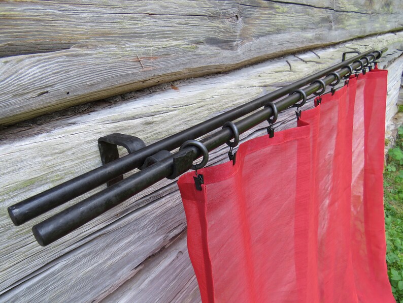 Op de afbeelding: Dubbele gordijnroede met zwarte metalen afwerking en rode, doorschijnende gordijnen. De roede is gemonteerd op een houten muur.