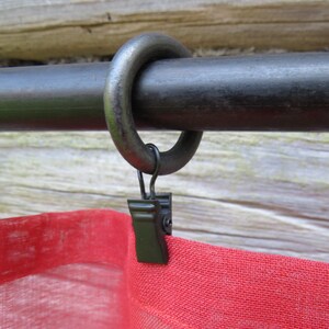 Op de afbeelding: Zwarte metalen gordijnroede met een zwarte metalen ring en een zwarte clip bevestigd aan een rood en wit gordijn.