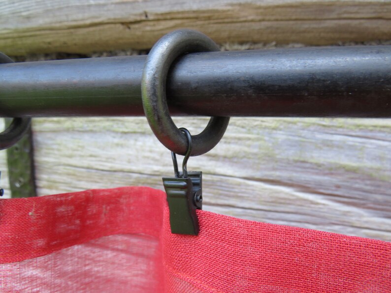 Op de afbeelding: Zwarte metalen gordijnroede met een zwarte metalen ring en een zwarte clip bevestigd aan een rood en wit gordijn.