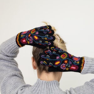 May include: A pair of black knit gloves with colorful embroidered floral designs. The gloves are worn on the hands of a person with their hair pulled back.