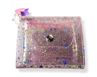May include: A clear, glitter-filled wallet with a pink hue. The wallet features a snap closure and a small charm with star and heart shapes. The glitter includes various colors and star shapes.