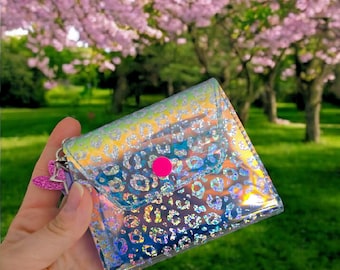 May include: A small iridescent clear purse with a leopard print design. The purse has a pink button closure and a silver chain.
