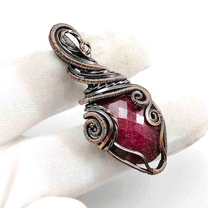 May include: A pendant featuring a faceted, deep red gemstone encased in intricate, oxidized copper wire wrapping. The metalwork consists of coiled and spiraled wire patterns that secure the stone, forming a decorative bail at the top.