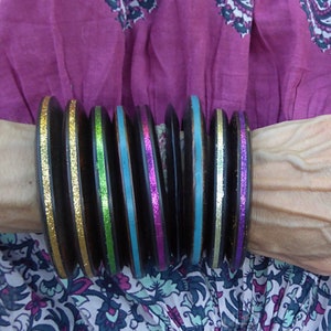 May include: A stack of black, round bangles with alternating glitter and metallic bands in gold, green, blue, and pink. The bracelets are stacked on a wrist, with a patterned fabric background.
