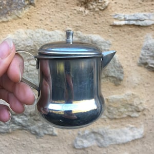 Peut inclure: Petite crémière ou saucière en acier inoxydable brillant, avec couvercle et anse. Le récipient a un corps arrondi et un bec verseur. L'anse est une simple boucle. Fond : mur en pierre brute.