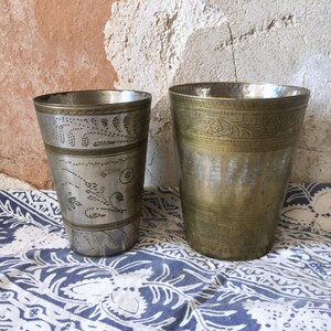 May include: Two antique metal drinking cups. One is silver-toned with etched floral designs and the other is gold-toned with etched floral designs. Both cups are cylindrical and sit on a blue and white patterned cloth.