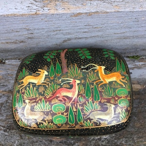 Op de afbeelding: Een rechthoekige, zwarte decoratieve doos met een geschilderde bosscène. De scène toont herten in geel, rood en wit, te midden van groene bladeren en bomen. De doos heeft een gouden rand en staat op een verweerd houten oppervlak.
