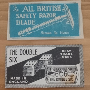 May include: Two rectangular vintage razor blade paper wrappers. The top wrapper is blue with white text reading The All British Safety Razor Blade and Second To None. The bottom wrapper is black and white with dice and the text The Double Six.