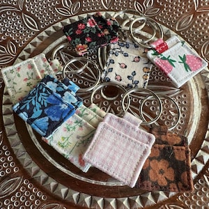 May include: A collection of square fabric keychains featuring various patterns including floral, gingham, and berry prints. Each keychain is attached to a silver metal split ring. The fabric squares are stitched with a small pocket-like detail on the front.