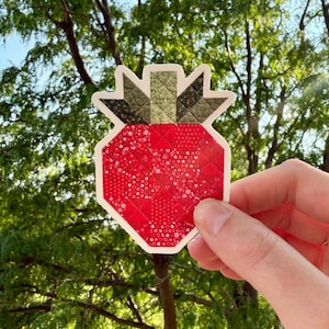 May include: A die-cut sticker shaped like a strawberry with a quilted texture. The main body is a vibrant red with a subtle white dot pattern, while the top leaves and stem are a dark olive green with a cross-hatched, quilted fabric design.