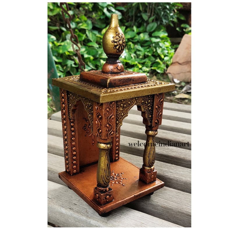 Puede incluir: Un peque&ntilde;o templo de madera tallado intrincadamente con un acabado dorado y cobre. El templo tiene una parte superior decorativa con un remate y un peque&ntilde;o arco. El templo est&aacute; sentado sobre una superficie de madera.