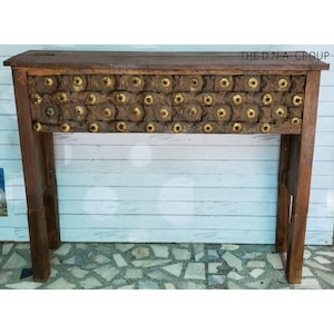 Puede incluir: Una mesa consola rectangular de madera con acabado marrón oscuro. El panel frontal presenta una fila horizontal de bloques de madera texturizados, cada uno adornado con un perno de metal color latón. El texto THE D.N.A. GROUP es visible arriba a la derecha.
