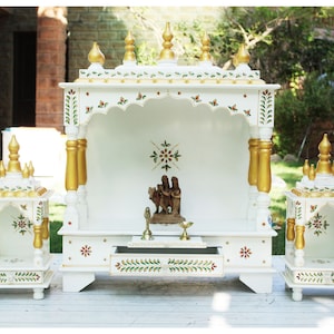 Può includere: Tempio in legno bianco con accenti dorati e intricati disegni floreali. Il tempio ha un piccolo cassetto e uno scaffale per statue o altri oggetti religiosi. Il tempio è decorato con motivi floreali verdi e rossi.
