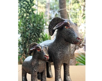 May include: Two ram figurines with a textured, dark metallic finish. Each ram features curved horns, pointed ears, and distinct orange eyes with red accents around the mouth and eyes. The figures stand on four legs, with one larger ram and one smaller ram.
