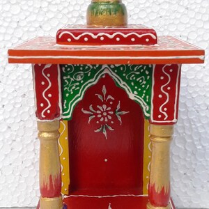 Puede incluir: Un peque&ntilde;o templo de madera pintado a mano con una base roja, acentos verdes y dorados y un remate dorado. El templo tiene un peque&ntilde;o nicho para una deidad o un &iacute;dolo.