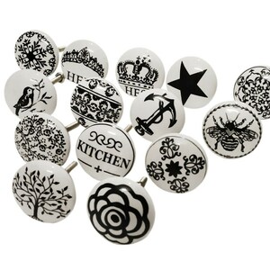 May include: Set of 12 white ceramic cabinet knobs with black designs. The designs include a crown, an anchor, a bee, a star, a bird, a tree, a rose, a snowflake, and various floral patterns. Some knobs have the word "Kitchen" written on them.
