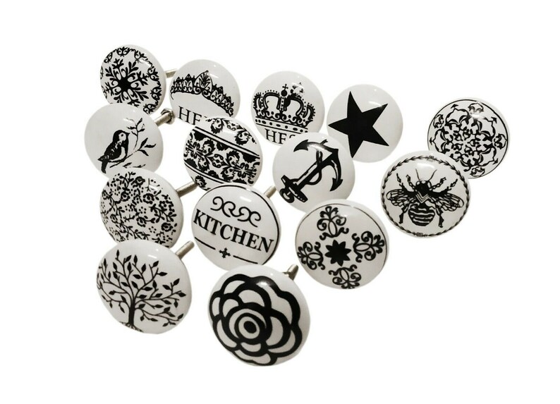 May include: Set of 12 white ceramic cabinet knobs with black designs. The designs include a crown, an anchor, a bee, a star, a bird, a tree, a rose, a snowflake, and various floral patterns. Some knobs have the word "Kitchen" written on them.