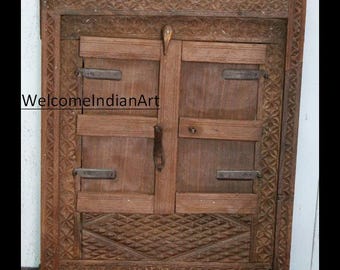 May include: A rectangular wooden window frame featuring intricate carved geometric patterns along the border and a diamond-shaped relief at the base. The frame includes two central shutters with dark metal hinges and a latch. Text reads WelcomeIndianArt.