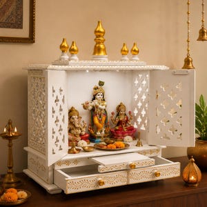 Könnte beinhalten: Ein weißer hölzerner Haustempel mit aufwendigen Gittertüren, goldfarbenen Akzenten und zwei Schubladen. Im Inneren befinden sich Statuen von Ganesha, Krishna und Lakshmi auf einem Regal, zusammen mit Ringelblumen und Messinggegenständen.