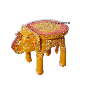 Puede incluir: Un taburete de elefante de madera pintado a mano con una parte superior redonda. El elefante es amarillo con diseños florales coloridos y la parte superior está decorada con un patrón de mandala rosa, amarillo y verde. El taburete es una pieza de mobiliario única y decorativa.