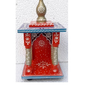 Puede incluir: Un peque&ntilde;o templo de madera rojo con un remate dorado en la parte superior. El templo tiene detalles pintados en blanco y acentos azules y morados. Est&aacute; decorado con motivos florales.
