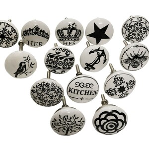 May include: Set of 14 white ceramic cabinet knobs with black and white designs. The designs include a bird, a crown, a star, a bee, a flower, a tree, an anchor, and various floral patterns. The knobs have a smooth, glossy finish.