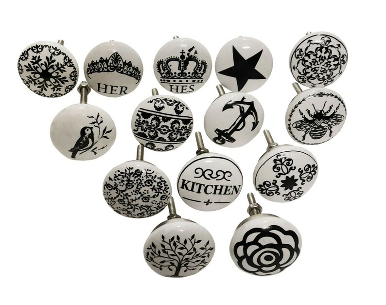 May include: Set of 14 white ceramic cabinet knobs with black and white designs. The designs include a bird, a crown, a star, a bee, a flower, a tree, an anchor, and various floral patterns. The knobs have a smooth, glossy finish.
