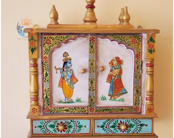 May include: A small, wooden, hand-painted shrine with two doors and a drawer. The doors are decorated with images of Hindu deities, Krishna and Radha, in colorful clothing. The shrine is painted in gold, green, blue, and red.