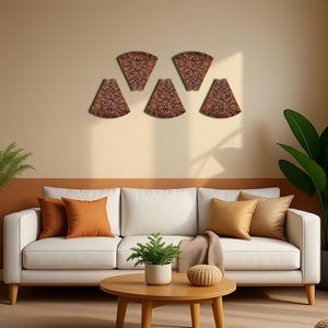 Könnte beinhalten: Fünf dekorative, braune, fächerförmige Wandkunstwerke mit aufwendigen floralen Mustern sind an einer beigefarbenen Wand angebracht. Die Stücke befinden sich über einem weißen Sofa mit orangefarbenen und goldenen Kissen. Ein Holztisch mit einer Pflanze und einer dekorativen Kugel steht vor dem Sofa.