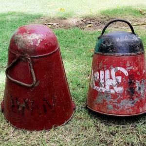 May include: Two vintage red metal buckets with black accents and white lettering. The bucket on the left has the word "SHIVAM" written on it. The bucket on the right has the word "LUCK" written on it.