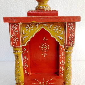 Puede incluir: Un peque&ntilde;o templo de madera pintado a mano con una base roja, acentos amarillos y dise&ntilde;os florales blancos. El templo tiene un techo puntiagudo y una peque&ntilde;a abertura en la parte delantera.