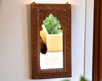May include: A rectangular wall mirror featuring a dark brown wooden frame with intricate hand-carved floral and leaf patterns. The mirror has an arched top opening and two small metal loops at the top edge for mounting purposes on a wall.