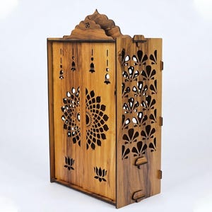 K&ouml;nnte beinhalten: Ein h&ouml;lzerner Heimtempel mit nat&uuml;rlicher Holzmaserung, aufwendigen lasergeschnittenen Lotusblumenmustern auf den Vordert&uuml;ren, Seitenteilen mit f&auml;cherf&ouml;rmigen Ausschnitten, Glockenmotiven und einem in den Bogen geschnitzten Om-Symbol.