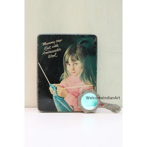 Può includere: Una scatola di latta vintage con una ragazza che fa la maglia e il testo "Mummy says Knit with Ambassador Wool". La scatola è accanto a uno specchio da mano d'argento con un vetro turchese.
