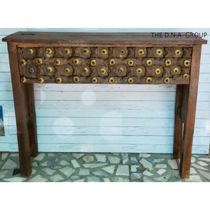 May include: A rustic wooden console table with a unique design featuring a decorative panel of metal studs and circles. The table has a dark brown finish and is supported by two sturdy legs.