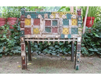May include: A colorful wooden cabinet with a decorative front panel featuring geometric shapes and a small door. The cabinet is painted in shades of red, green, yellow, and blue, and has a distressed finish. It sits on four legs with decorative carvings.