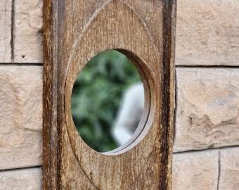 Puede incluir: Un espejo de pared rectangular con un marco de madera desgastada de color marrón oscuro que presenta un diseño ovalado puntiagudo y elevado alrededor de un centro de vidrio circular. La veta de la madera es visible y tiene un gancho de metal arriba.