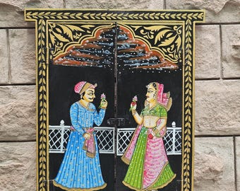 May include: A rectangular black wooden wall hanging shaped like a window with two doors. The surface features a hand-painted scene of two figures in traditional attire holding flowers, framed by a gold leaf-style border and decorative arch motifs.