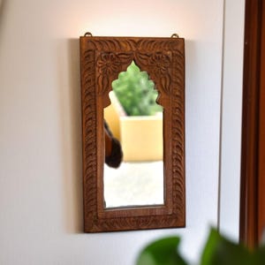 Op de afbeelding: Een rechthoekige houten spiegel met een gewelfde bovenkant en gedetailleerde bloemsnijwerk. De spiegel is omlijst in donkerbruin hout en hangt aan een witte muur. Het ontwerp doet denken aan de Indiase of Marokkaanse stijl.