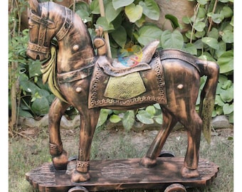 May include: A bronze-colored horse statue mounted on a rectangular wheeled platform. The horse features detailed harness work, a saddle with a textured gold-toned panel, and a flowing tail. The entire structure is finished with a weathered patina.
