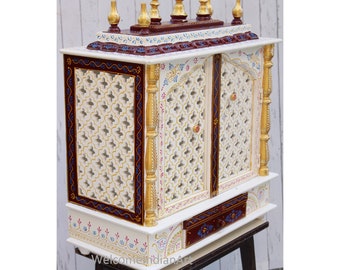 May include: White wooden temple with intricate carvings and gold accents. The temple has two doors with a lattice design and a small drawer below. The temple is sitting on a wooden stand.