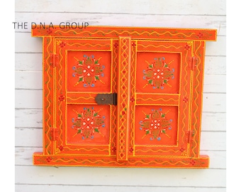 May include: A bright orange wooden window frame with two hinged shutters, featuring hand-painted floral patterns in green, blue, and white. The frame includes a dark metal latch and the text THE D.N.A. GROUP printed in black on the upper left corner.