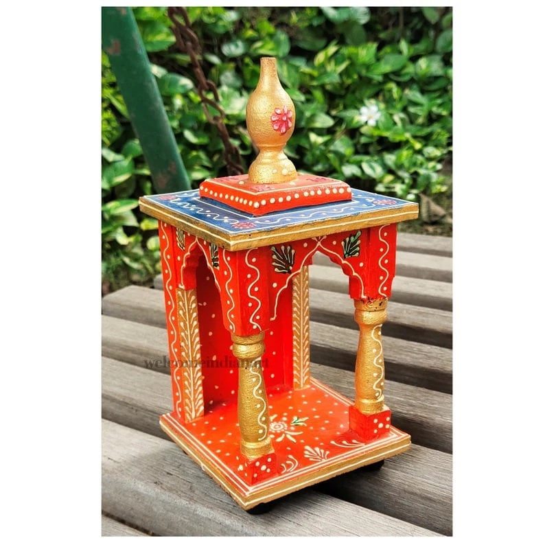 Puede incluir: Un peque&ntilde;o templo de madera pintado a mano con una base roja, detalles dorados y una parte superior azul. El templo tiene un arco decorativo y un peque&ntilde;o remate dorado en la parte superior.