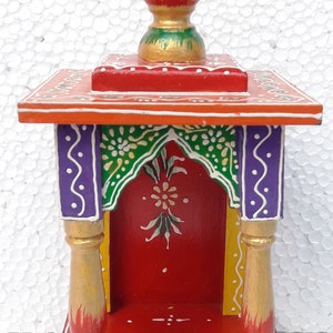 Puede incluir: Un peque&ntilde;o templo de madera pintado a mano con una base roja, acentos verdes y morados y un remate pintado en oro. El templo tiene una peque&ntilde;a abertura en la parte delantera para colocar una estatua u otros objetos religiosos.